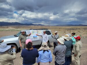 Two men presenting graphics and maps on a poster hanging from the side of a truck to a group of collaborative researchers from Peru in south-central Nevada.
